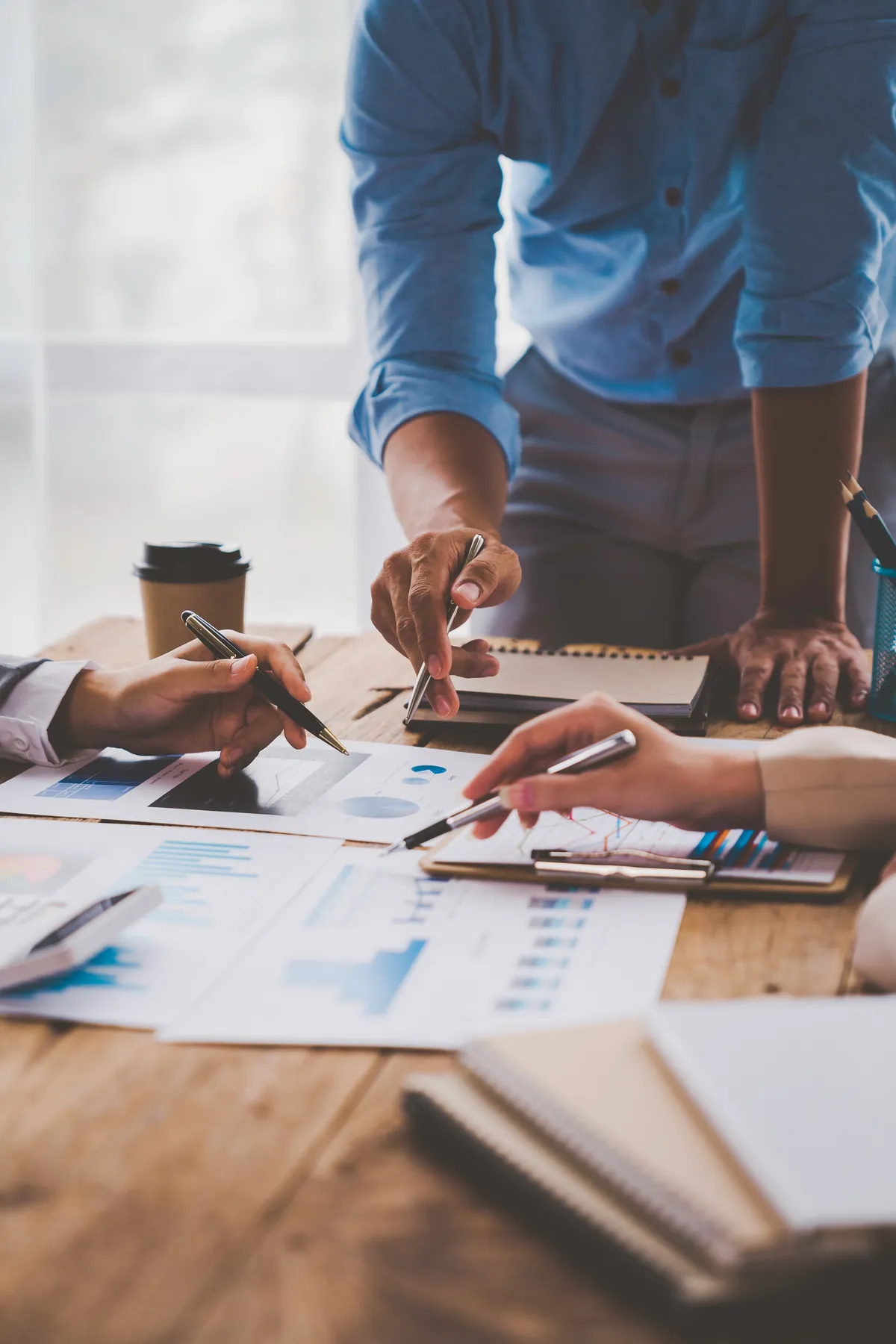 Colleagues point to charts and notes spread across a wooden table during a planning session