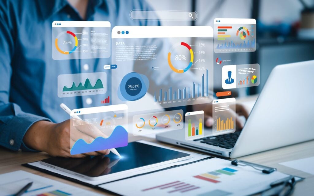 A person working on a tablet and laptop as charts and data dashboards float above the desk